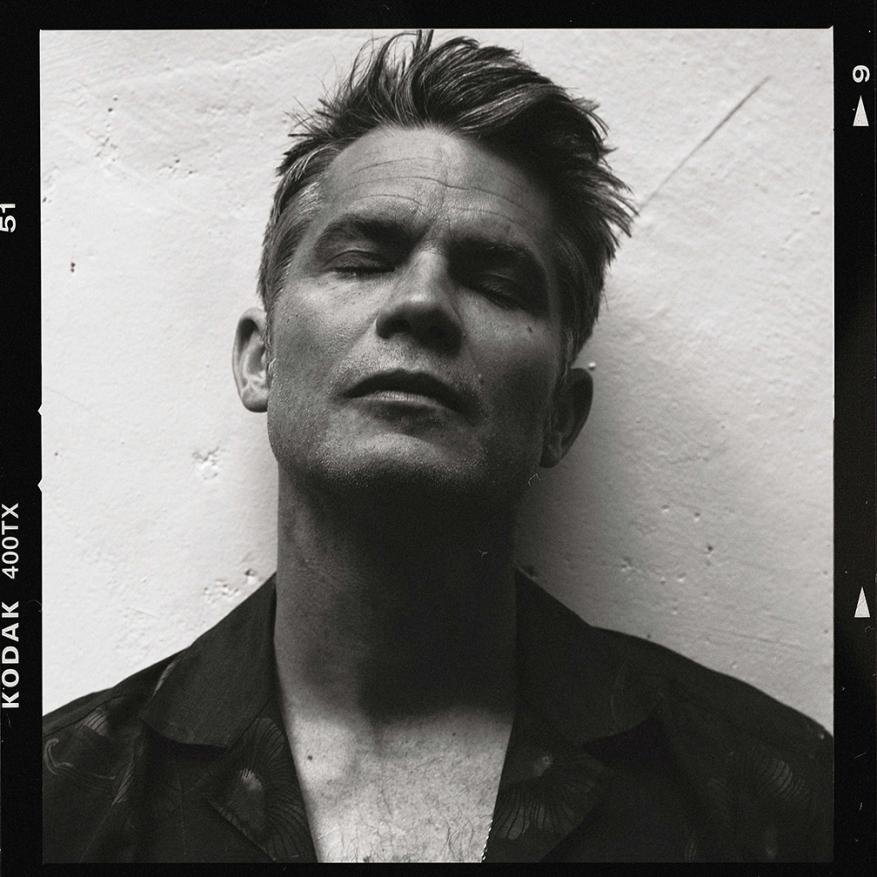 Black-and-white film portrait of an adult male with short hair and eyes closed, wearing a floral shirt in front of a light-colored wall.