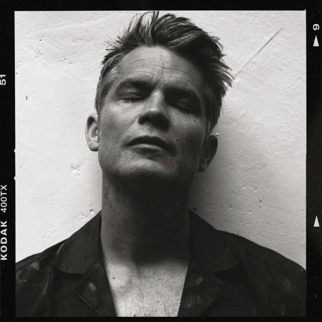 Black-and-white film portrait of an adult male with short hair and eyes closed, wearing a floral shirt in front of a light-colored wall.