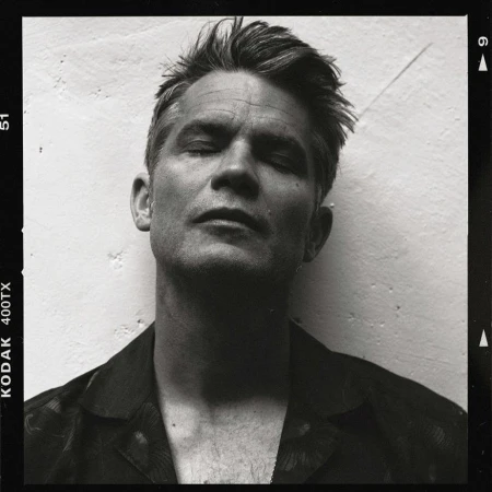 Black-and-white film portrait of an adult male with short hair and eyes closed, wearing a floral shirt in front of a light-colored wall.