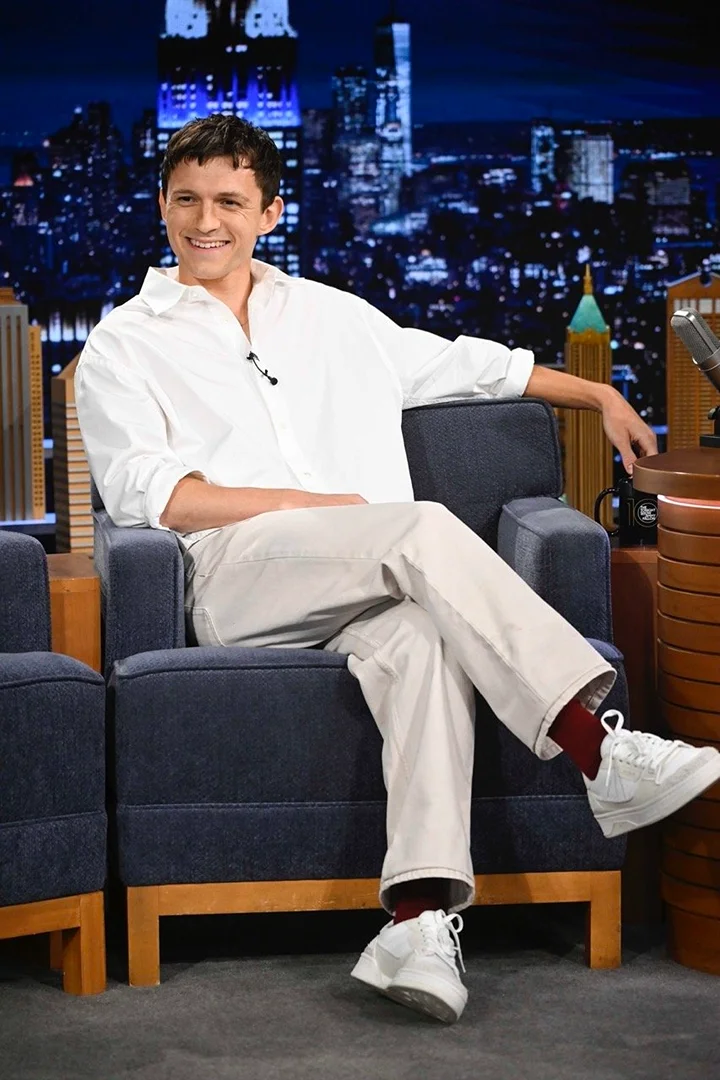 Smiling man in a white shirt sits in a blue chair on a talk show set, with a backdrop of city lights at night.