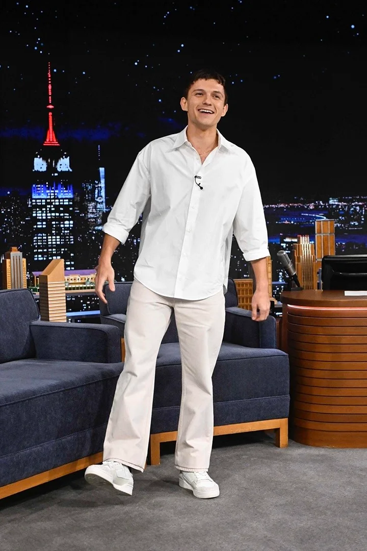 Tom Holland stands on stage in a white shirt and light-beige pants in front of a city backdrop and studio couch setting for a show.