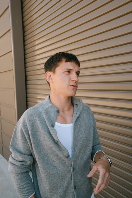 Portrait of a young adult male wearing a grey cardigan and a white t-shirt standing outdoors against a beige roll-up door.
