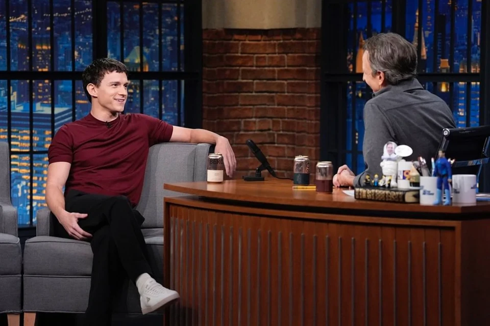 Two men sit in a television studio. One man in a maroon shirt is interviewing the other at a wooden desk with city views behind them.