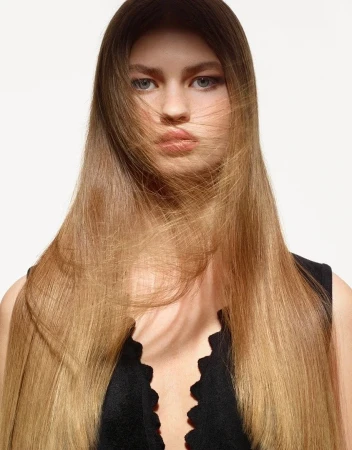 Close-up portrait of a woman with long brown hair blowing across her face wearing a black scalloped top against a white background
