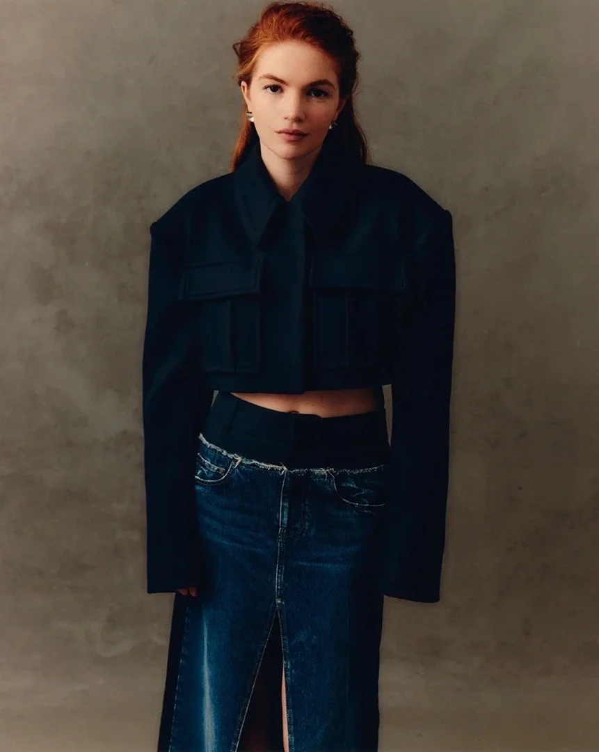 Fashion portrait of a redhead woman wearing a dark cropped jacket and a blue denim skirt with a front slit against a grey background.
