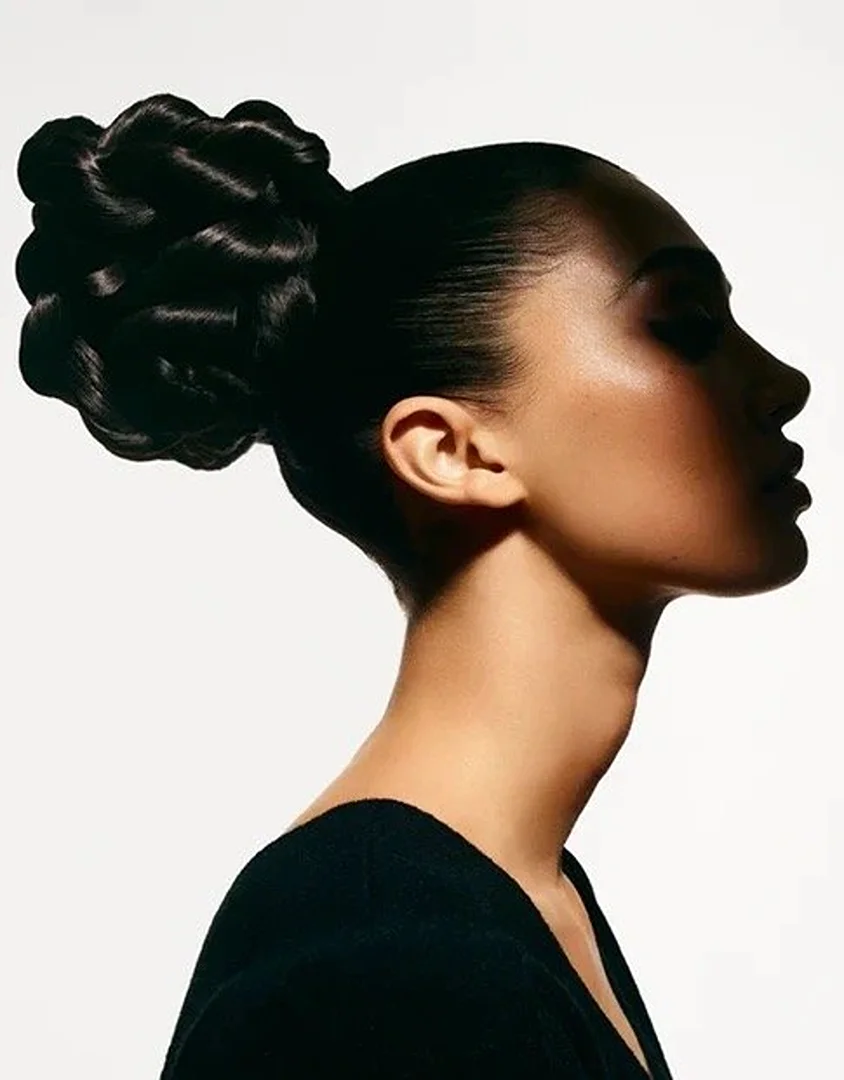 Profile shot of a dark-haired woman with a high bun hairstyle, wearing a black top, set against a bright white studio backdrop.