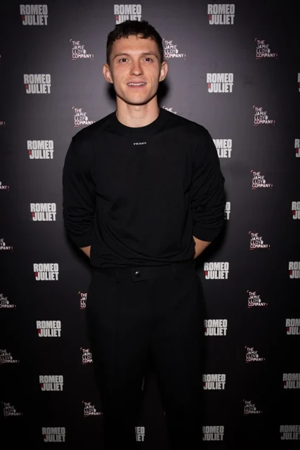 A smiling adult male poses in front of a Romeo and Juliet promotional backdrop, wearing a long-sleeved black shirt and black pants indoors.