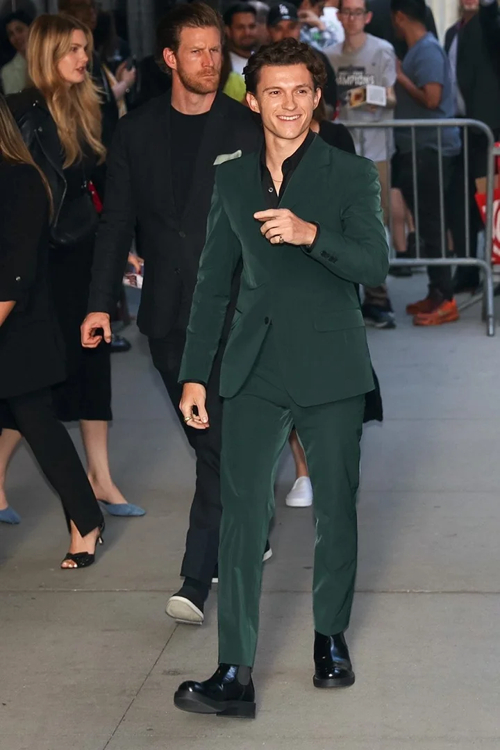 Tom Holland poses in an emerald suit on a city street. A group of people walks behind him on the sidewalk. He smiles at the camera.