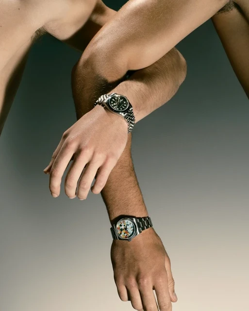 Close-up of two arms wearing wristwatches, one with a metal band and dark face, the other with a lighter face, against a grey backdrop.
