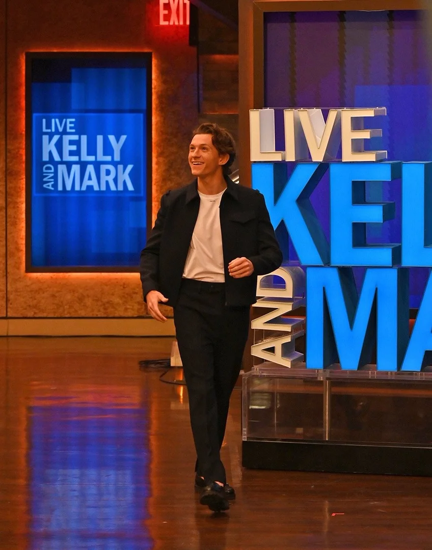 Tom Holland walking on the set of Live with Kelly and Mark, wearing a black jacket and pants with a white t-shirt.