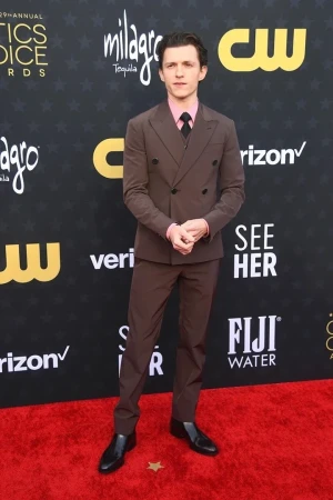 Tom Holland on the red carpet at the Critics Choice Awards, wearing a brown suit with a pink shirt and black shoes. Full shot.