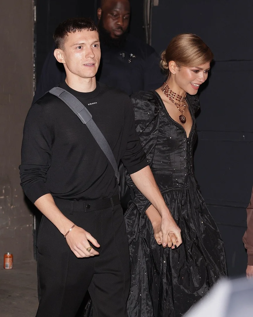 Tom Holland and Zendaya hold hands while walking with a security guard in the background. Zendaya wears a sparkling dress.