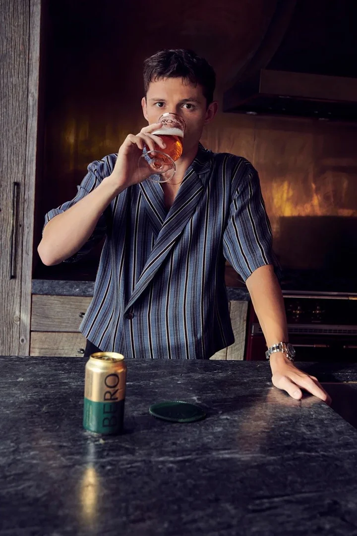 Adult man indoors drinking beer from a glass in front of an open beer can, wearing a striped short sleeve shirt, at counter.