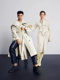 Man and woman posed against a light grey wall with dark flooring, wearing cream trench coat and knit dress styled by Crystalle Cox.