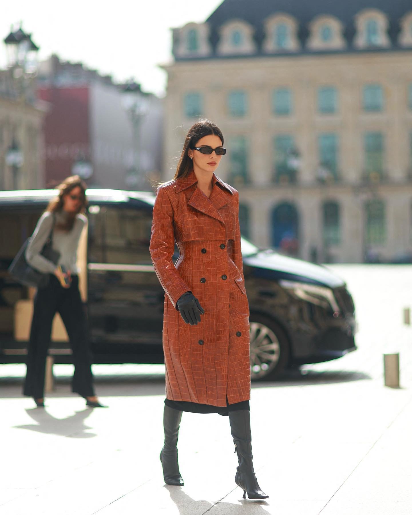 A woman in a patterned coat and boots stands on a city street with muted architecture, a vehicle, and another woman visible in the background.
