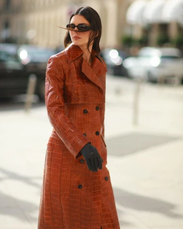 A woman with sunglasses poses in a long, brown, crocodile-patterned coat with black gloves, standing on a city street with cars and buildings.