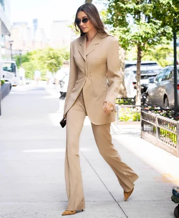 An adult female with sunglasses walks on a sidewalk wearing a beige suit with flared pants and pointed-toe shoes in an urban environment.