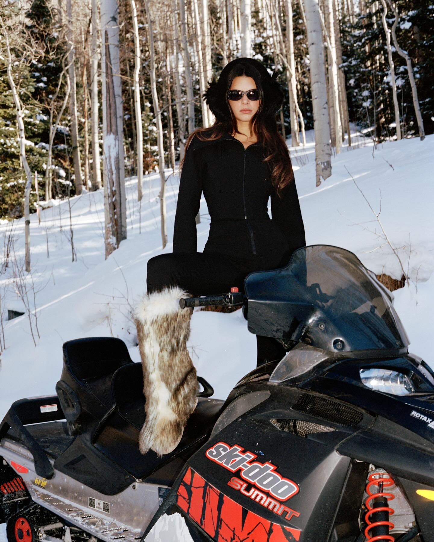 Woman in a black snowsuit poses on a Ski-Doo snowmobile in a snowy forest with tall, thin trees, wearing furry boots and sunglasses.