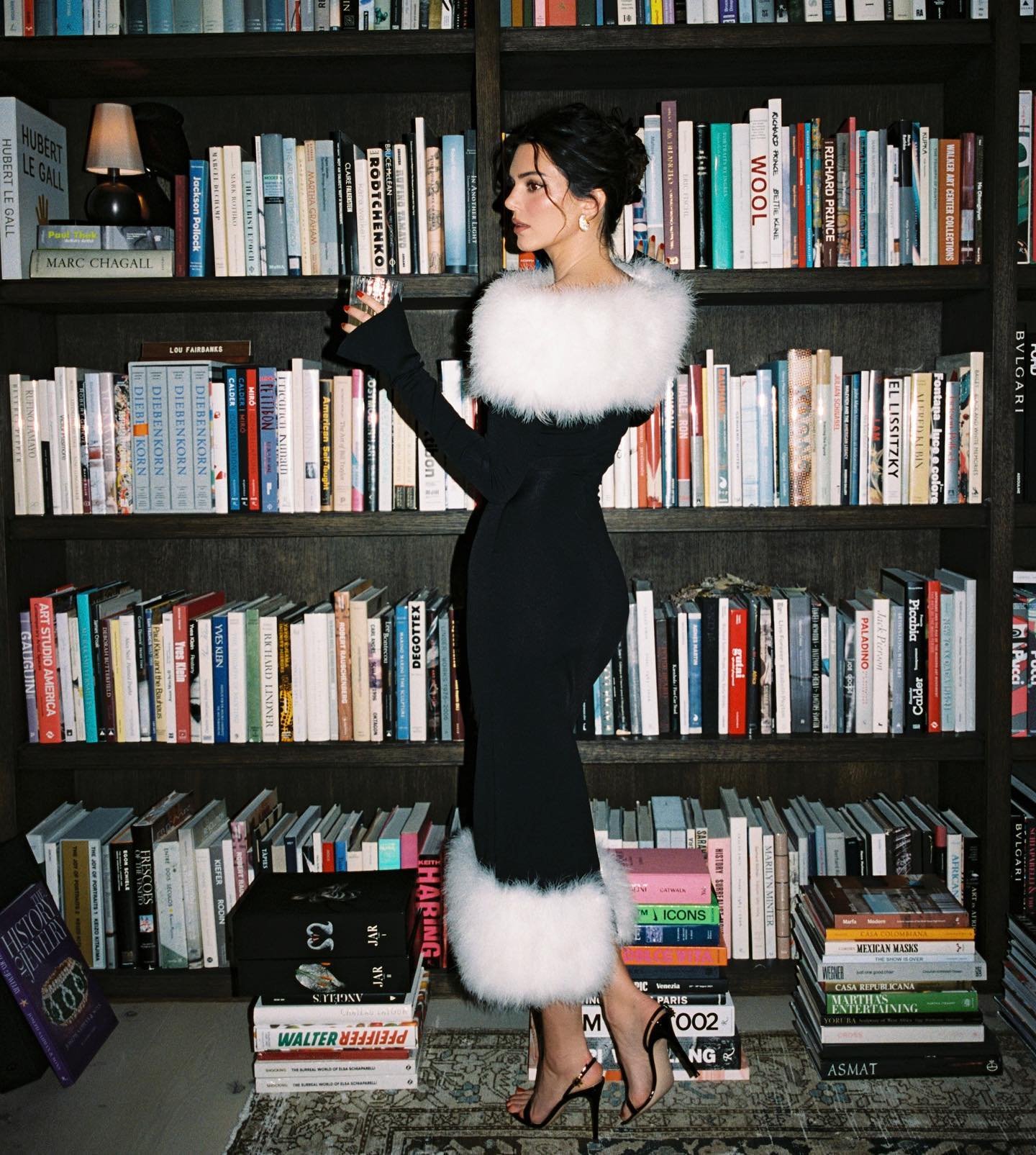 Woman in a stylish black dress with white faux fur trim poses in front of a bookcase filled with books, creating a fashionable portrait.