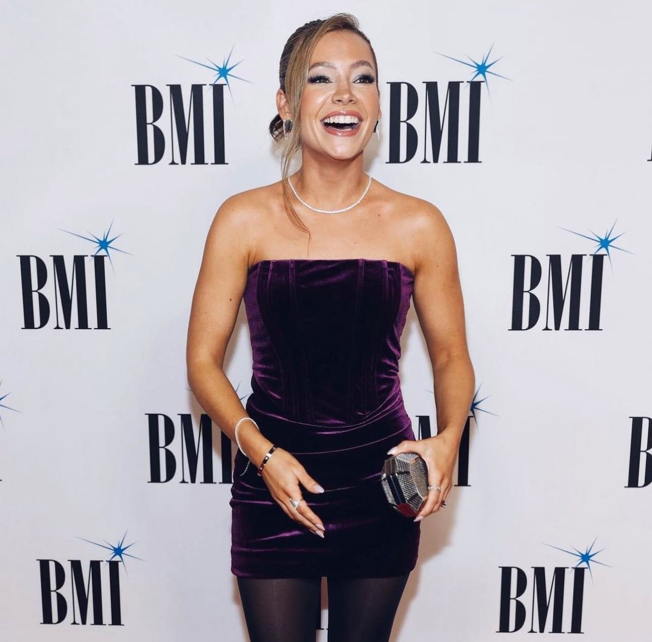 A cheerful woman in a strapless purple velvet dress smiles and poses on a red carpet at a BMI event with logo backdrop.