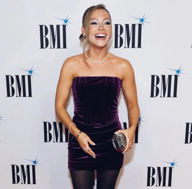 A cheerful woman in a strapless purple velvet dress smiles and poses on a red carpet at a BMI event with logo backdrop.