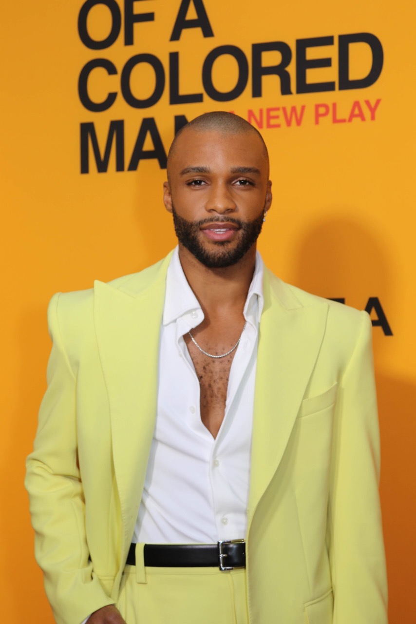 Portrait of a handsome, bald, African American man wearing a yellow suit and white button-down shirt in front of a yellow and black poster.
