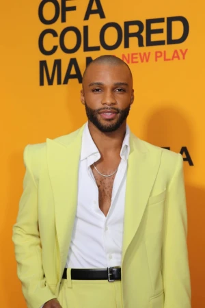 Portrait of a handsome, bald, African American man wearing a yellow suit and white button-down shirt in front of a yellow and black poster.
