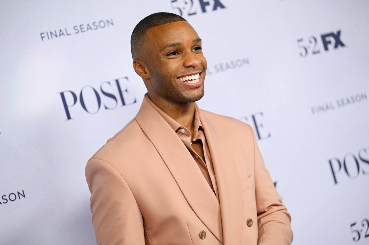 Portrait of an African-American man smiling in a light pink suit in front of a Pose final season backdrop. Formal wear, red carpet.