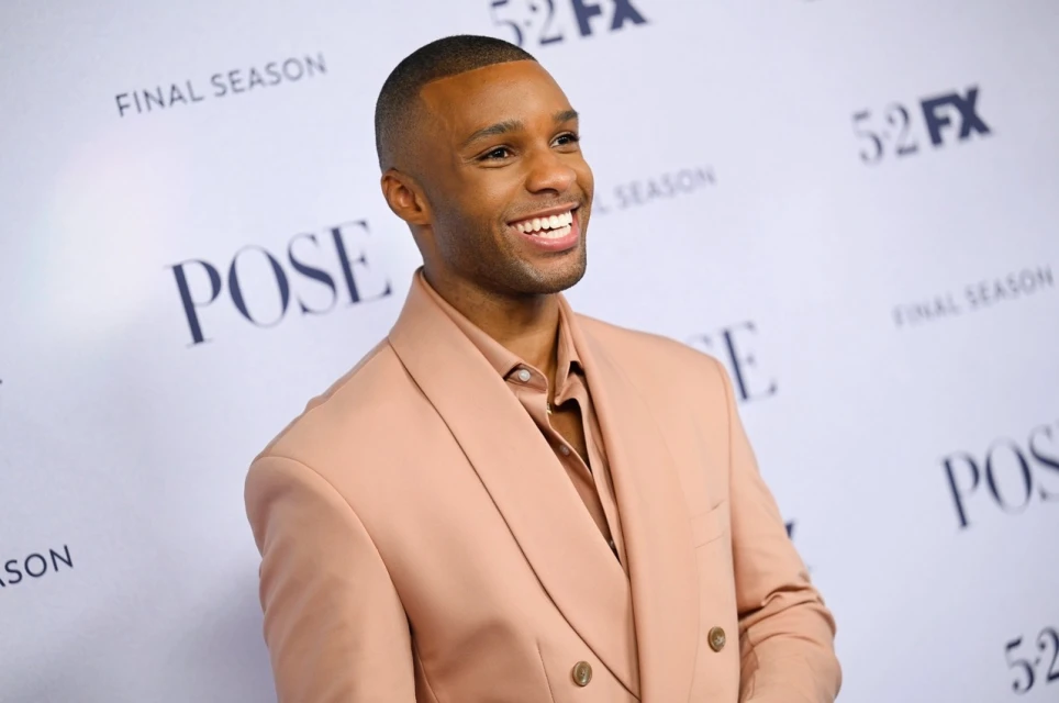 Portrait of an African-American man smiling in a light pink suit in front of a Pose final season backdrop. Formal wear, red carpet.