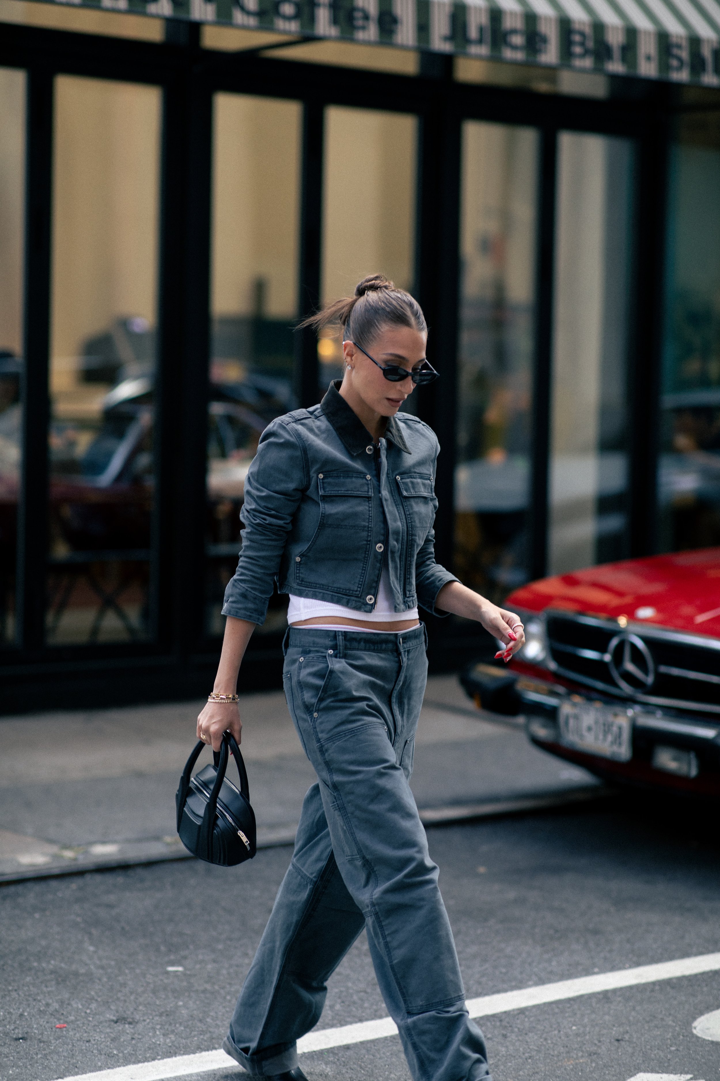 Adult female fashion model wearing a gray denim jacket and pants, black handbag, and sunglasses walking on city street.