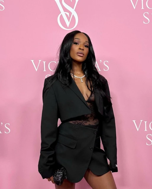 Elegant woman in a black blazer and lace top poses against a branded light-pink backdrop, accessorized with a necklace and clutch.
