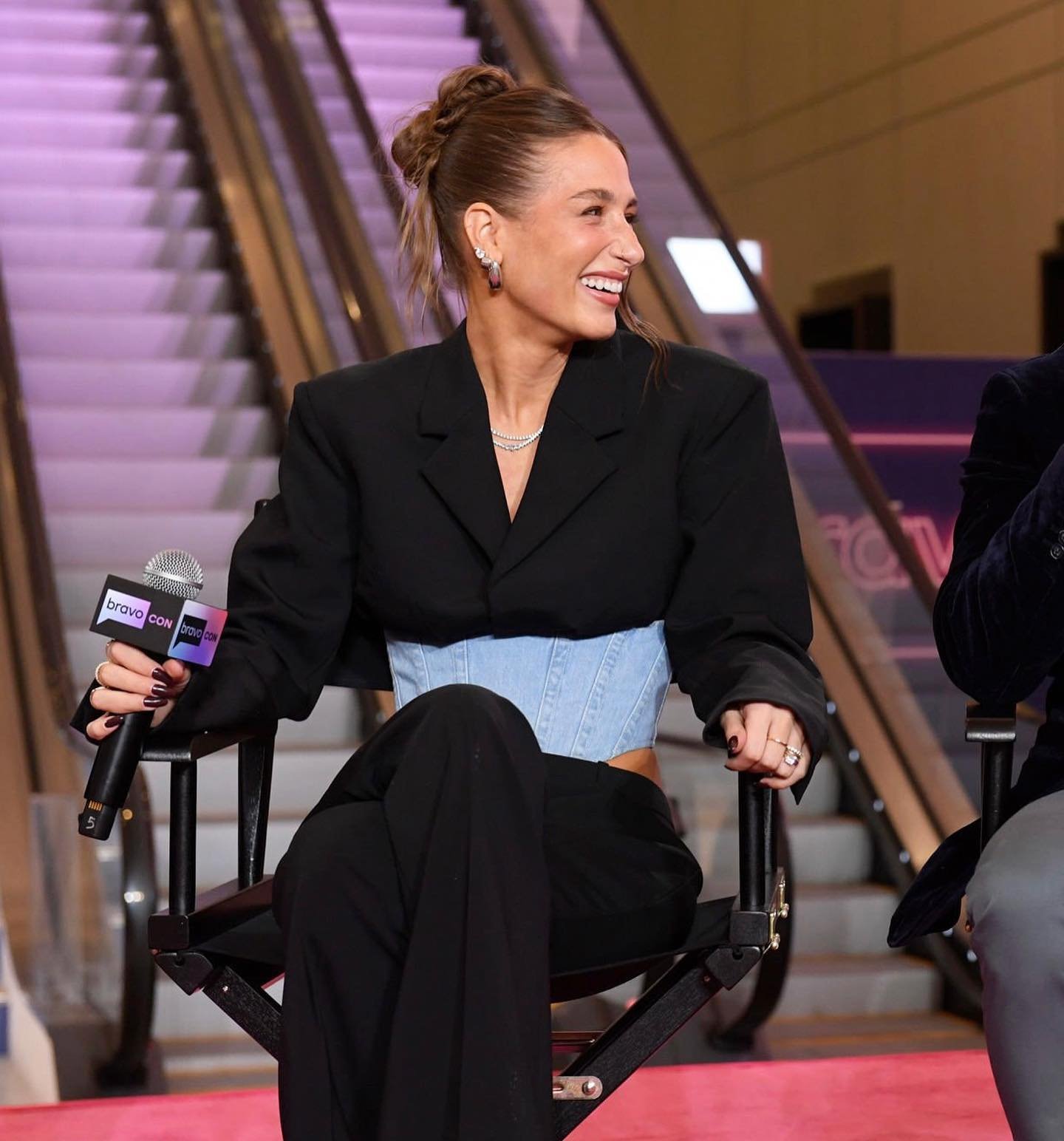 Smiling woman in a black blazer and blue corset sits in a chair on a pink carpet, holding a microphone with an escalator in the background.