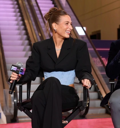 Smiling woman in a black blazer and blue corset sits in a chair on a pink carpet, holding a microphone with an escalator in the background.