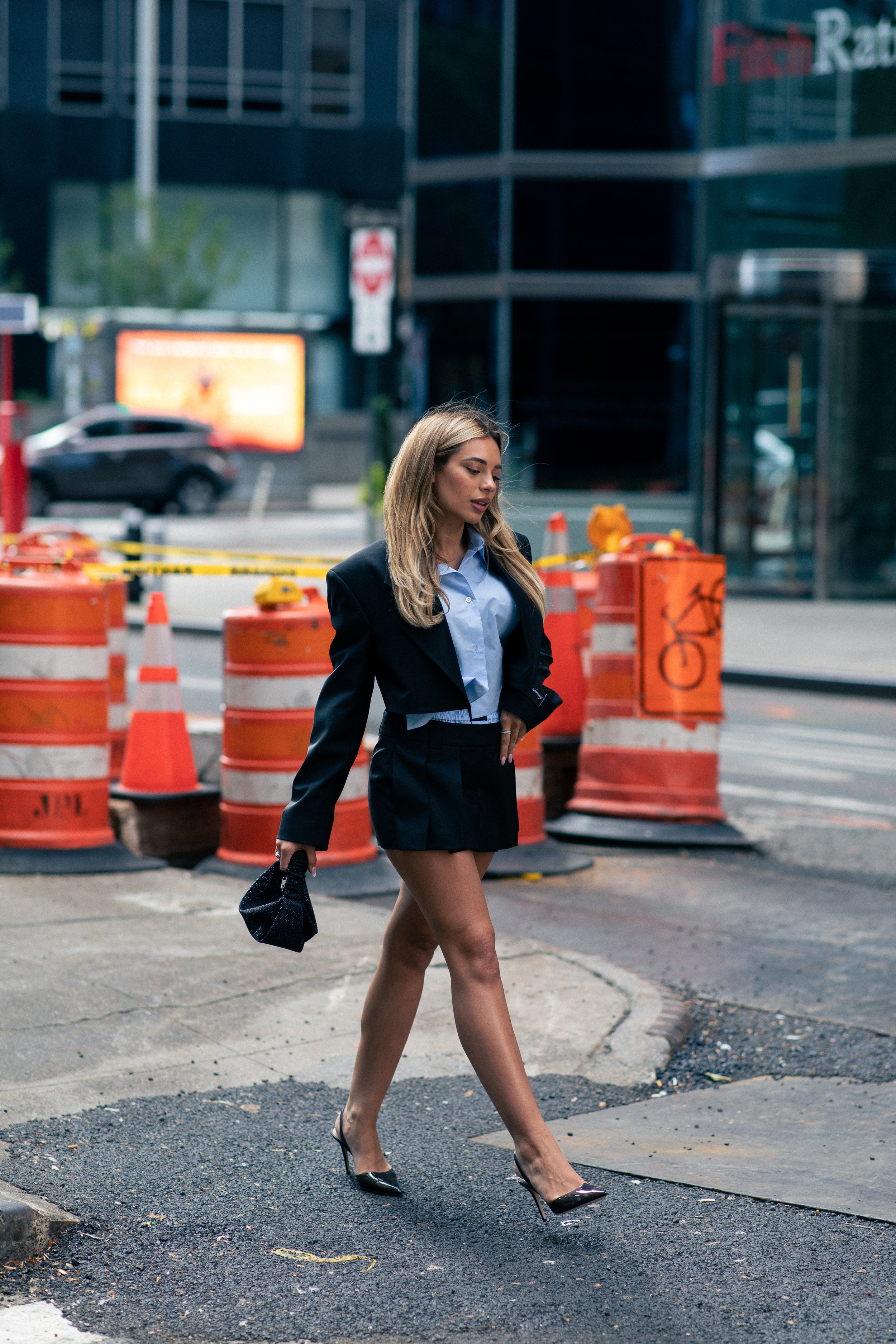 Woman in fashionable short black skirt suit walks down city street with traffic barrels, modern office buildings in the background.