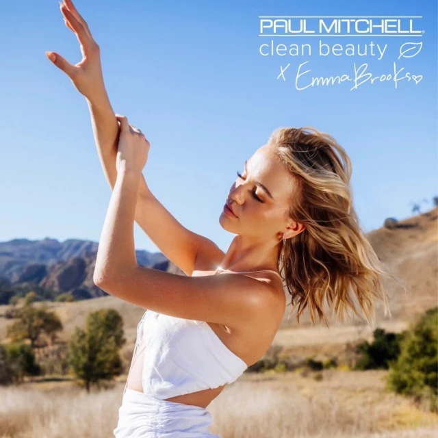 Model with arms raised poses in a field with a mountain backdrop wearing a white outfit, outdoor shot with branding text overlayed.