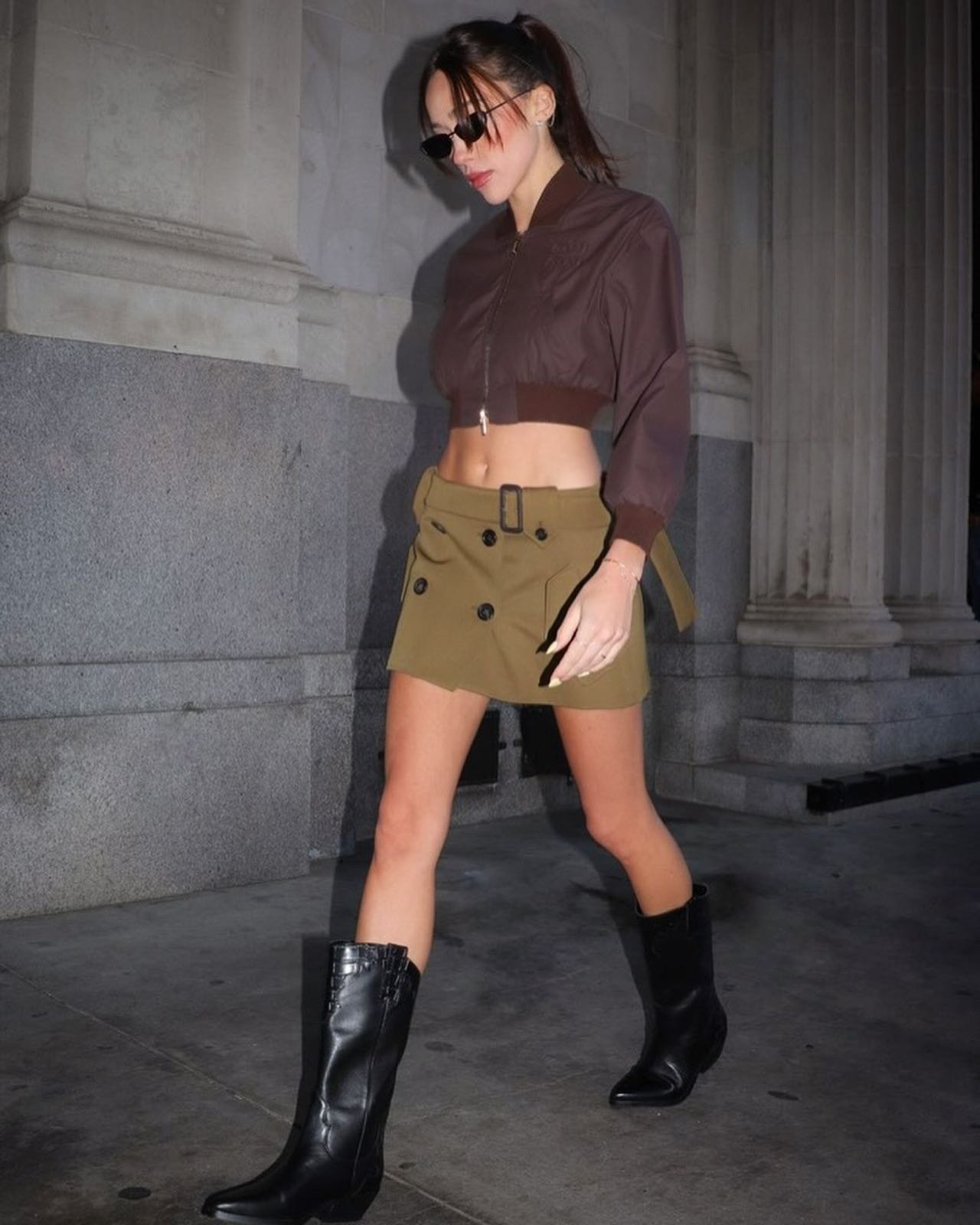 Stylish young woman in a brown bomber jacket and olive mini skirt walks on an urban street wearing sunglasses and black cowboy boots.