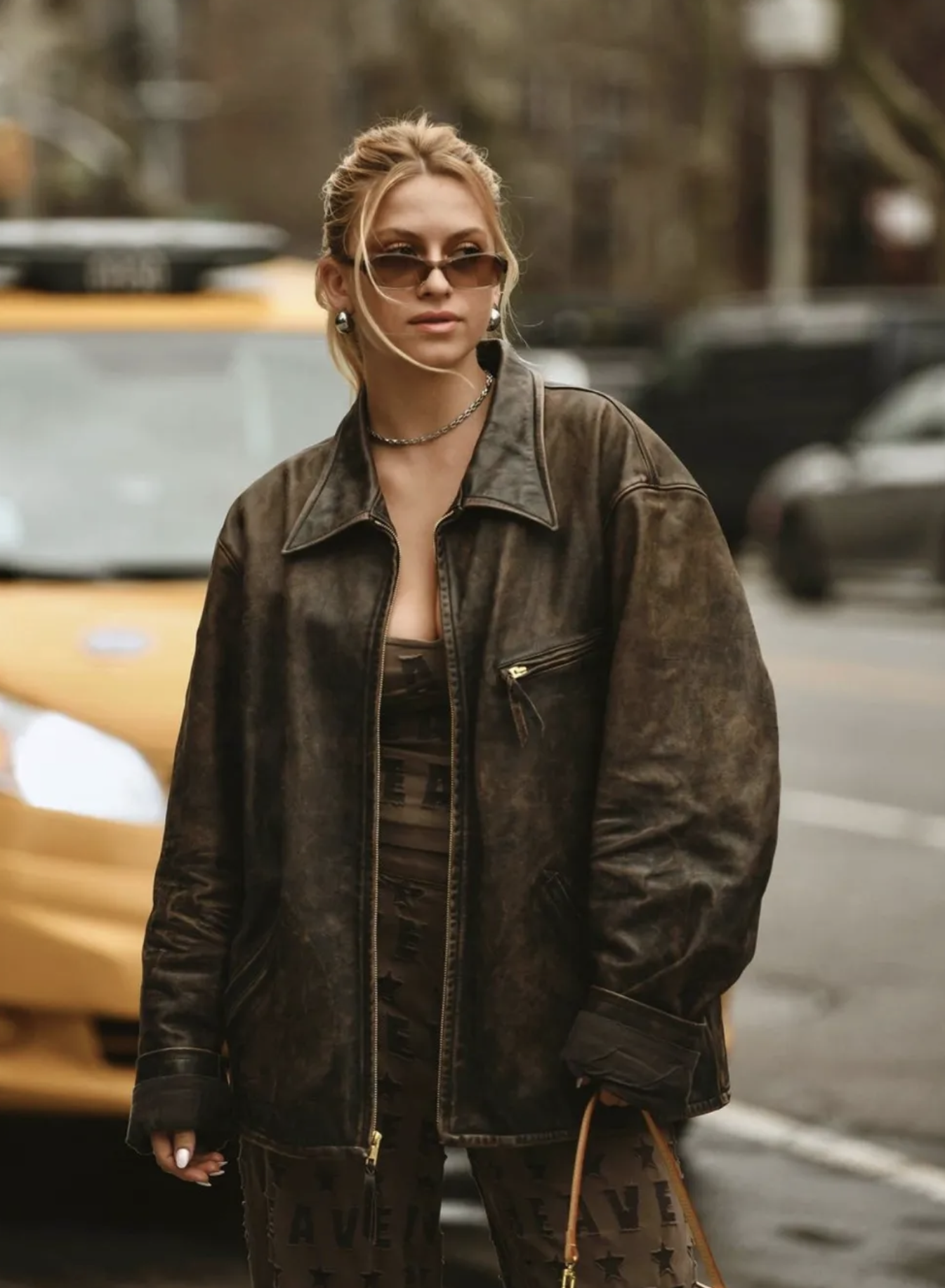 Stylish adult female in brown leather jacket and patterned jumpsuit stands on city street with yellow taxi in background.