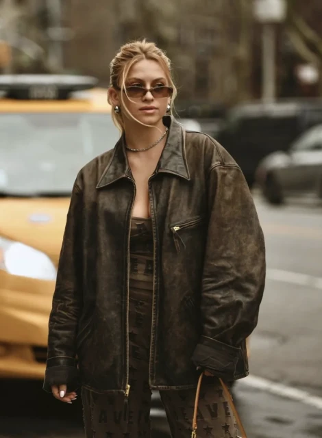 Stylish adult female in brown leather jacket and patterned jumpsuit stands on city street with yellow taxi in background.