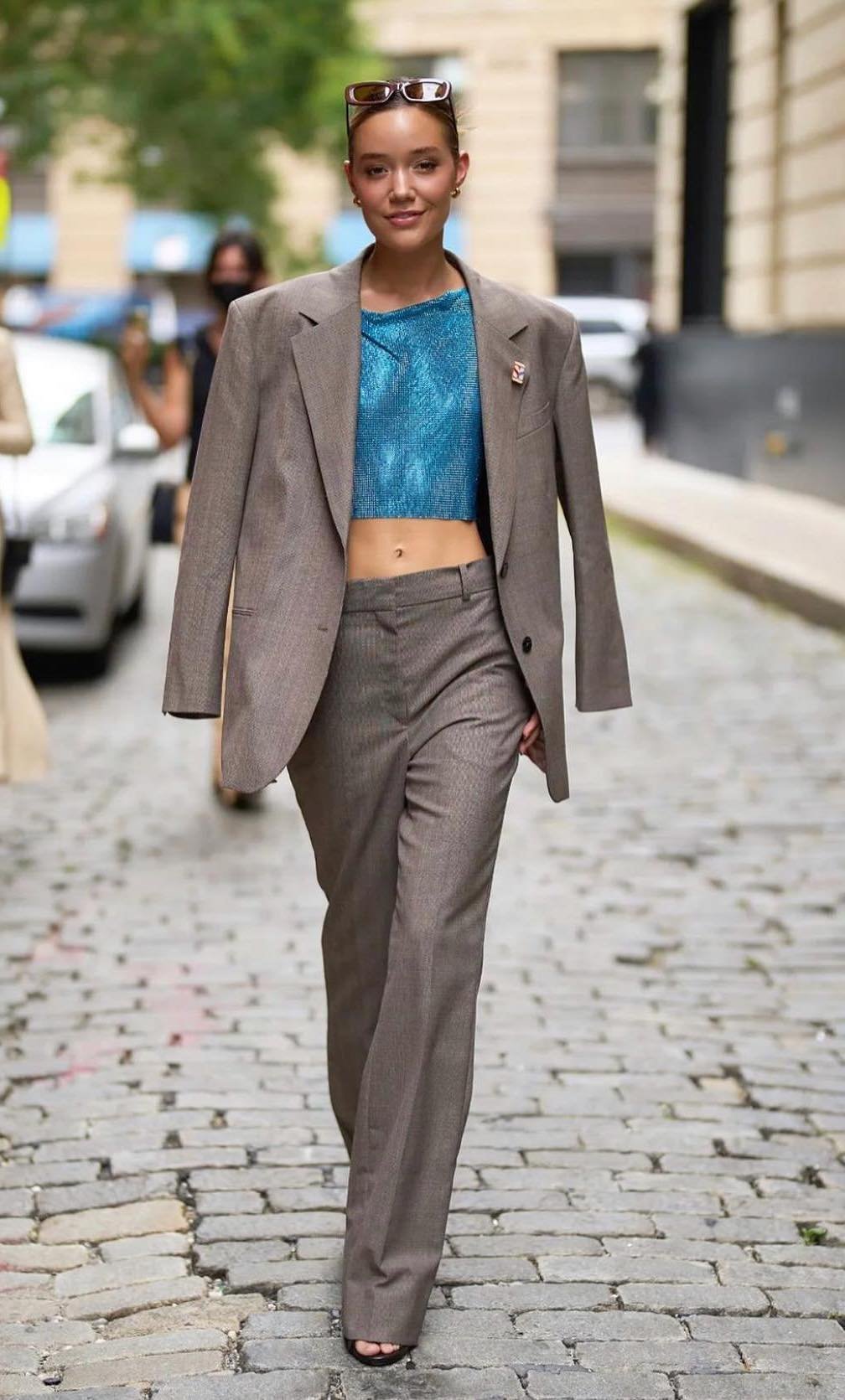 A woman in a grey suit stands on a cobblestone street wearing sunglasses and a blue top, posing in an outdoor urban setting.