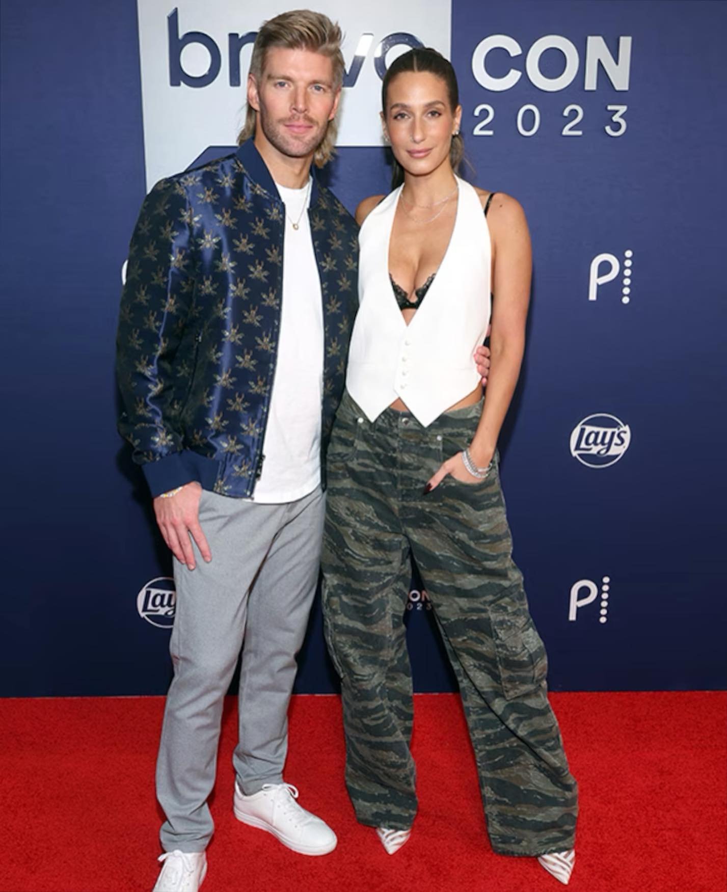 BravoCon 2023: A blonde man in a patterned jacket poses with a brunette woman in camouflage pants on a red carpet at a promotional event.