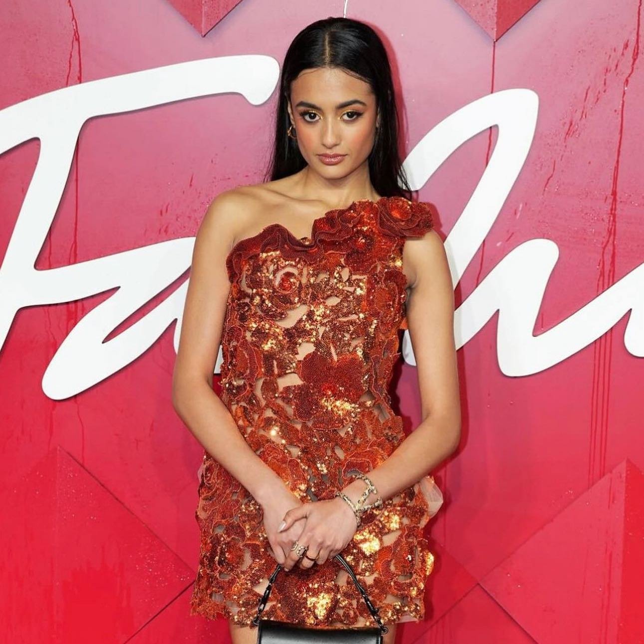 Woman in a sequined, rose-detailed, one-shoulder short dress at the Fashion Awards, smiles in front of a bright red backdrop.