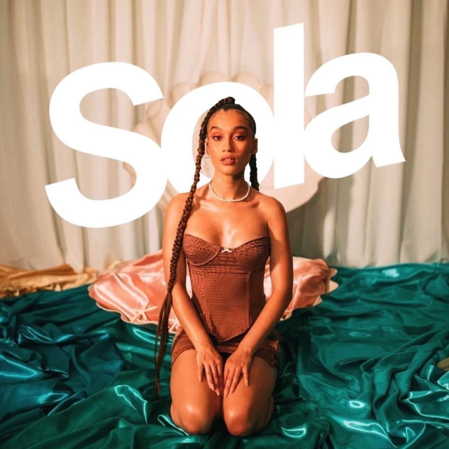 Young woman poses kneeling on satin fabric in a brown corset and mini skirt, pearl necklace, and box braids in a studio setting.