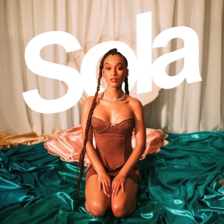 Young woman poses kneeling on satin fabric in a brown corset and mini skirt, pearl necklace, and box braids in a studio setting.