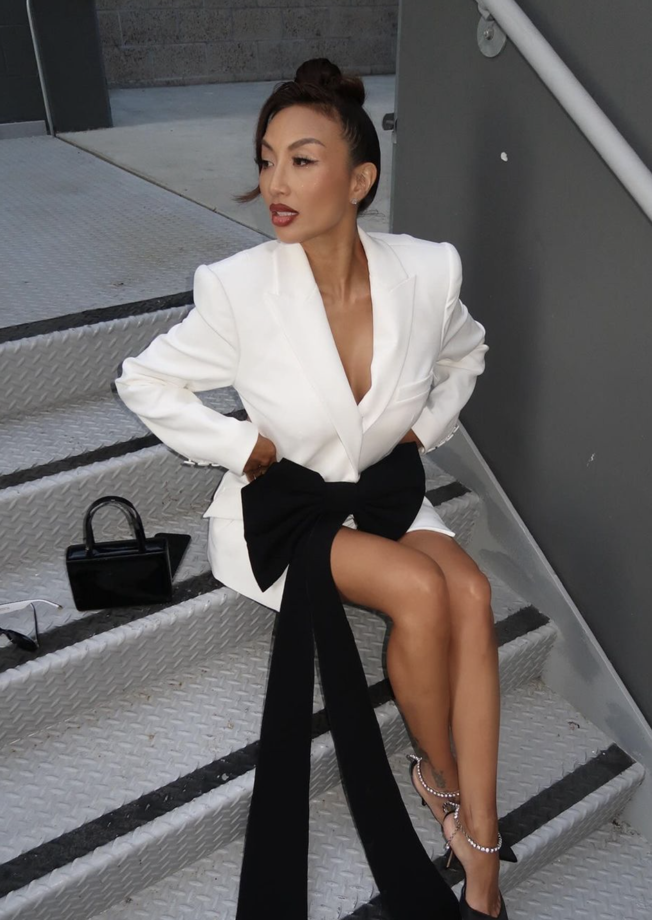 Elegant adult female in white blazer dress with black bow sitting on metal steps with a black purse and black high heels.