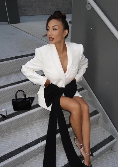 Elegant adult female in white blazer dress with black bow sitting on metal steps with a black purse and black high heels.