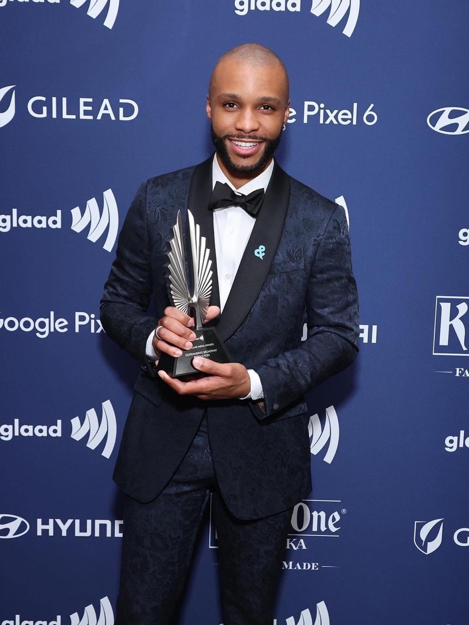 Well-dressed adult male holding a GLAAD award. He's wearing a dark floral suit, bow tie, and smiling at an awards ceremony event indoors.