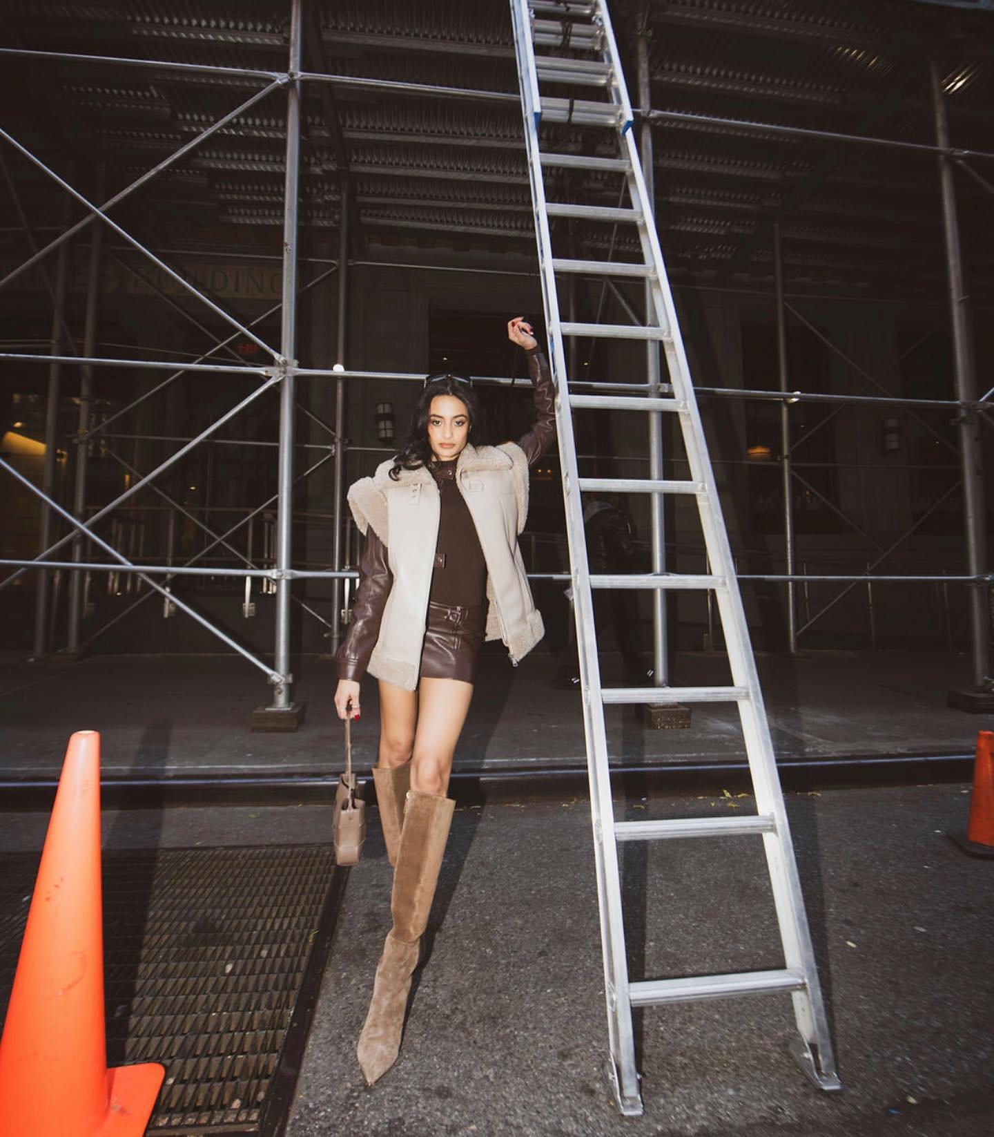 A woman in a brown dress and boots stands next to scaffolding and a ladder, holding a small bag and raising one arm up.