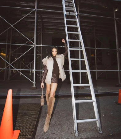 A woman in a brown dress and boots stands next to scaffolding and a ladder, holding a small bag and raising one arm up.