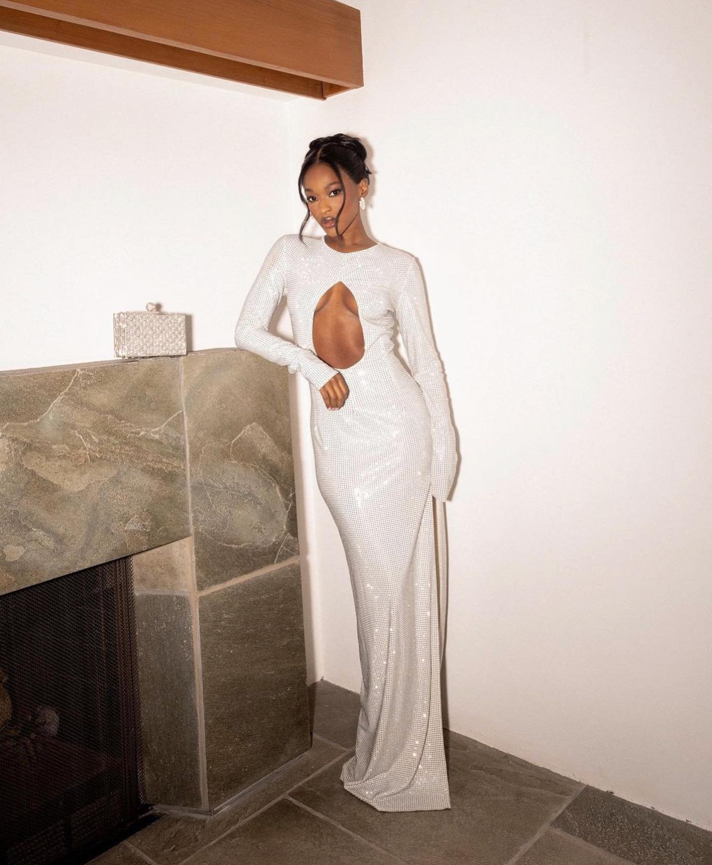 Glamorous adult female fashion model poses in a long-sleeve, cut-out white gown in an indoor setting with a fireplace and clutch.