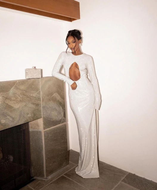 Glamorous adult female fashion model poses in a long-sleeve, cut-out white gown in an indoor setting with a fireplace and clutch.